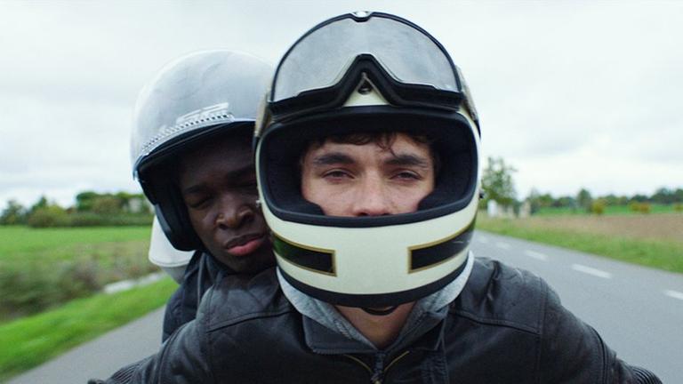 Die beiden Schauspieler Fionn Whitehead und Stéphane Bak sitzen mit Helmen auf einem Motorrad. Die beiden Schauspieler Fionn Whitehead und Stéphane Bak sitzen mit Helmen auf einem Motorrad.