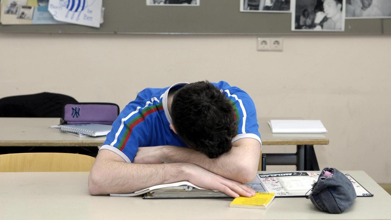 Ein Schüler sitzt im Klassenzimmer, den Kopf auf die Arme gestützt. Ein Schüler sitzt im Klassenzimmer, den Kopf auf die Arme gestützt.