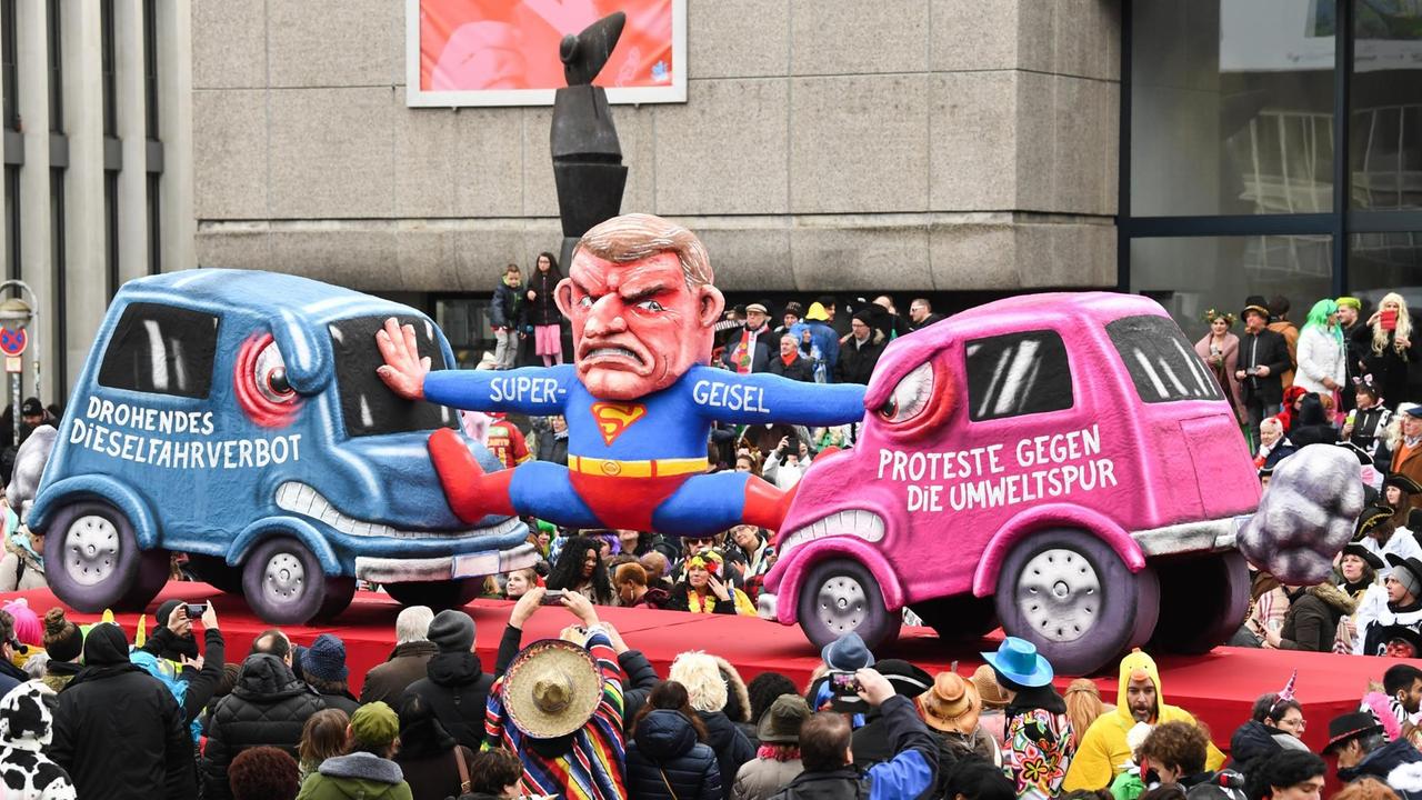 Beim Rosenmontagszug in Düsseldorf fährt ein Motivwagen mit zwei Autoattrappen durch die Menge. Auf einem steht: Drohendes Dieselfahrverbot. Auf dem anderen: Proteste gegen die Umweltspur. Dazwischen eine Figur im Supermankostüm, die den Düsseldorfer OB Thomas Geisel darstellen soll.