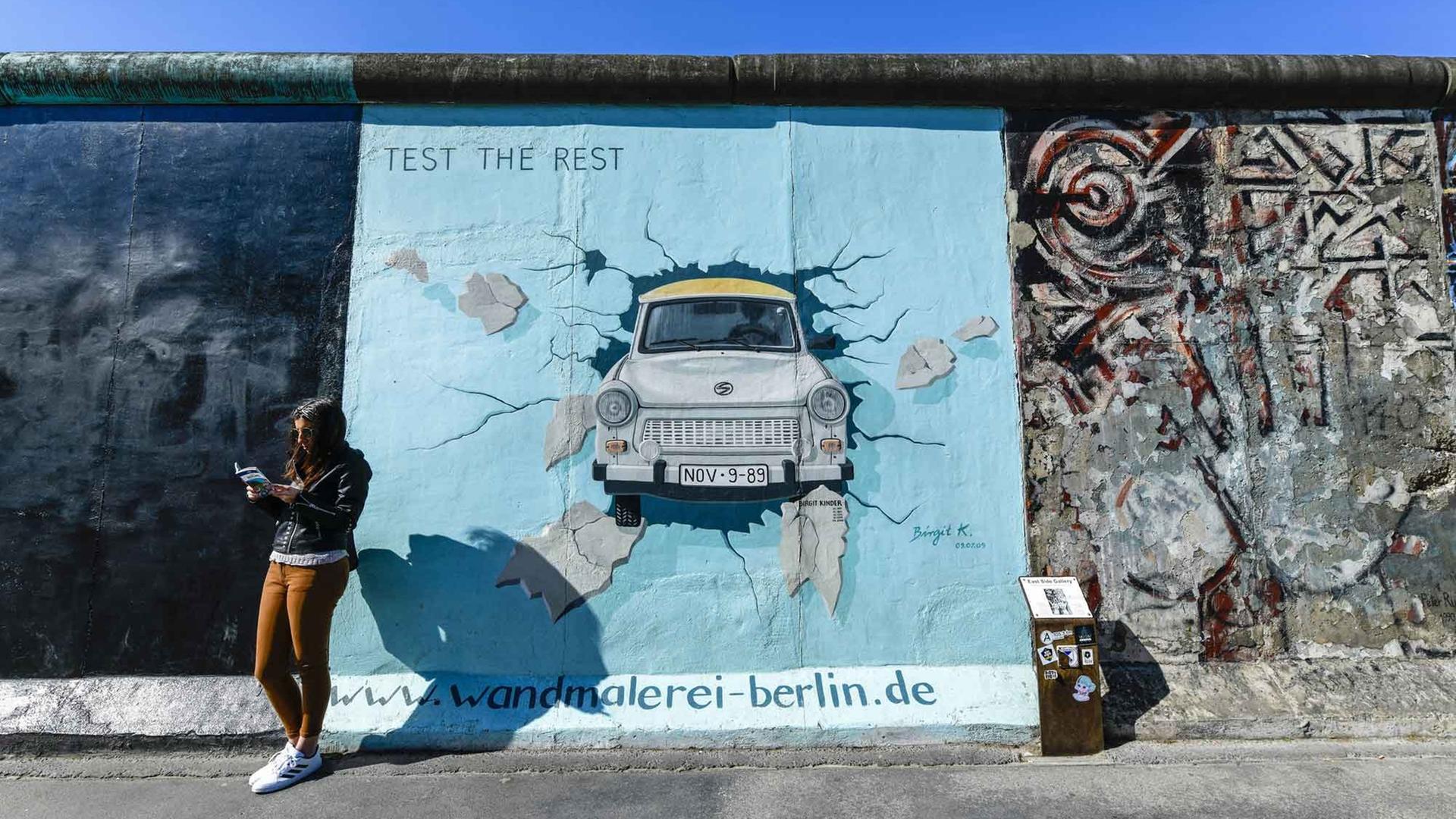 Eine junge Frau steht lesend vor der Berliner Mauer. Darauf ist eine Wandmalerei: ein Trabi, der die Mauer durchbricht.