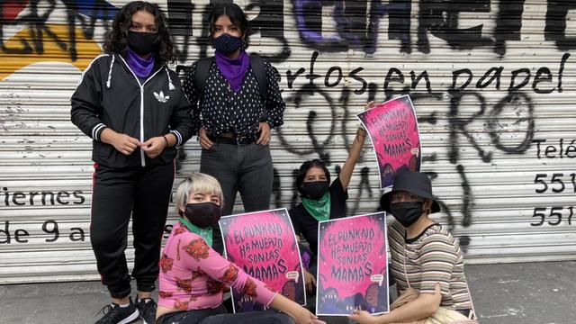 Fünf junge Frauen mit Masken und Plakaten stehen und sitzen auf der Straße vor einem Gebäude mit geschlossenen Rollos, die mit Graffiti besprüht sind.