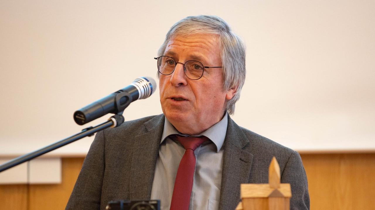 Ein bebrillter Mann im grauen Anzug spricht in ein Mikrofon. Ein bebrillter Mann im grauen Anzug spricht in ein Mikrofon.
