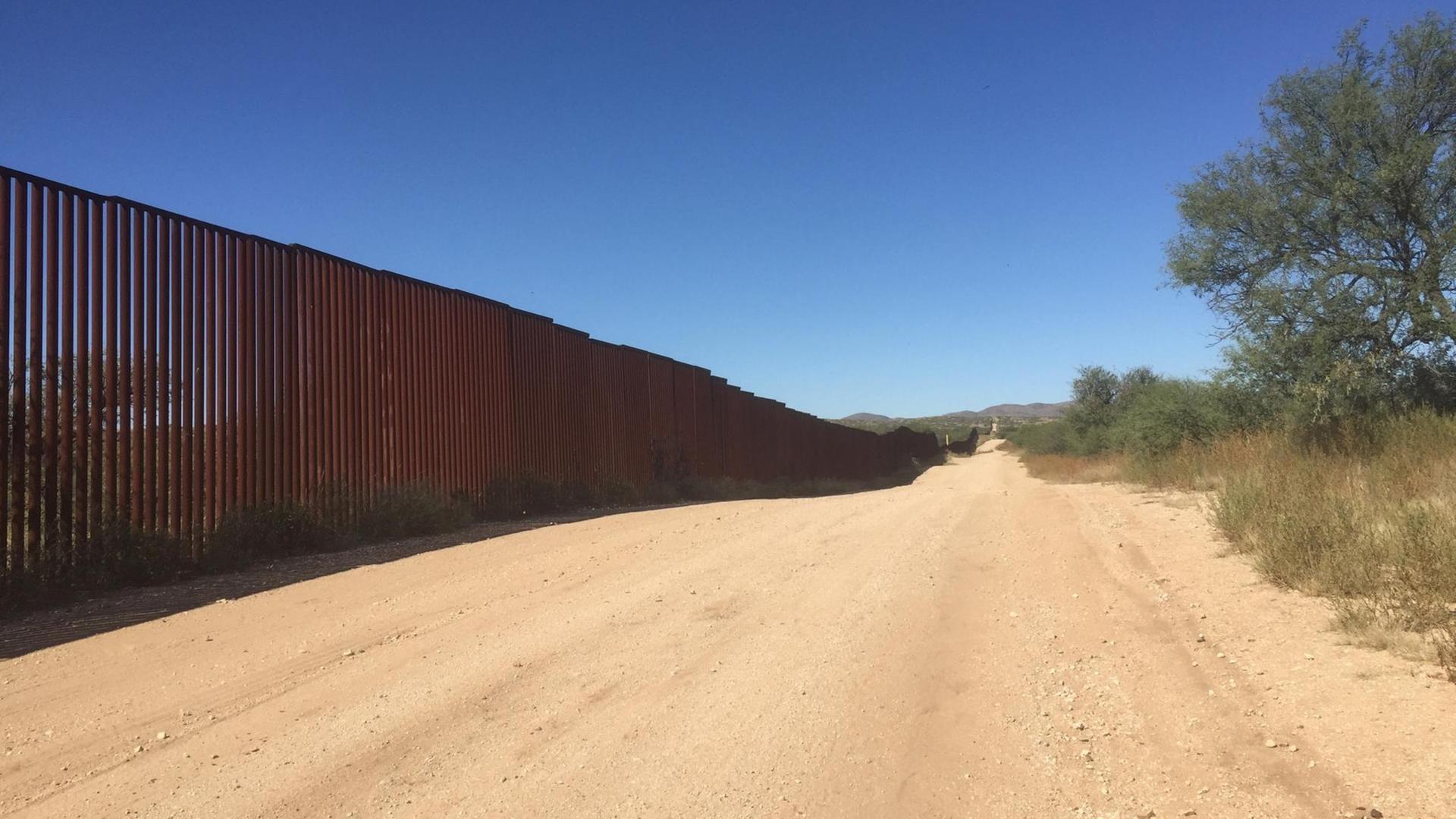 Am linken Bildrand sieht man den Grenzzaun zwischen Mexiko und den USA. In der Bildmitte verläuft ein sandiger Weg und am rechten Bildrand sind karge Bäume und Büsche zu sehen. Am linken Bildrand sieht man den Grenzzaun zwischen Mexiko und den USA. In der Bildmitte verläuft ein sandiger Weg und am rechten Bildrand sind karge Bäume und Büsche zu sehen.