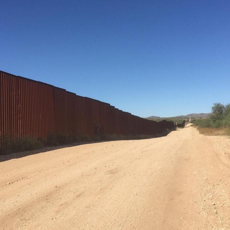 Am linken Bildrand sieht man den Grenzzaun zwischen Mexiko und den USA. In der Bildmitte verläuft ein sandiger Weg und am rechten Bildrand sind karge Bäume und Büsche zu sehen. 