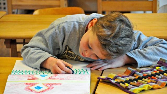 Ein circa achtjähriger blonder Junge hängt über ein Bild gebeugt und malt eifrig mit Kreiden. Er befindet sich in einer Waldorfschule.
