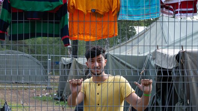 Ein junger Mann steht in einem Flüchtlingslager hinter einem Zaun.