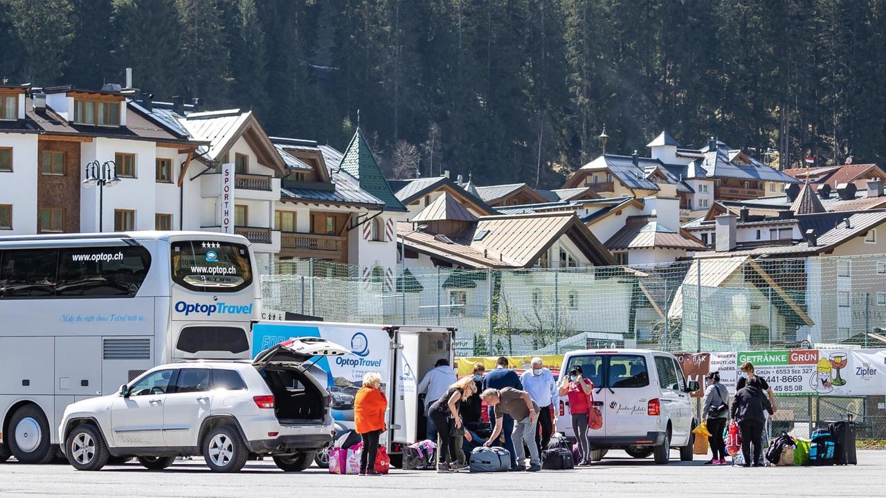 Eine Gruppe von Menschen ist mit Gepäck an einem Parkplatz um Autos und einen Bus versammelt, im Hintergrund ist die Häuswersilhouette von Ischgl zu sehen. Eine Gruppe von Menschen ist mit Gepäck an einem Parkplatz um Autos und einen Bus versammelt, im Hintergrund ist die Häuswersilhouette von Ischgl zu sehen.