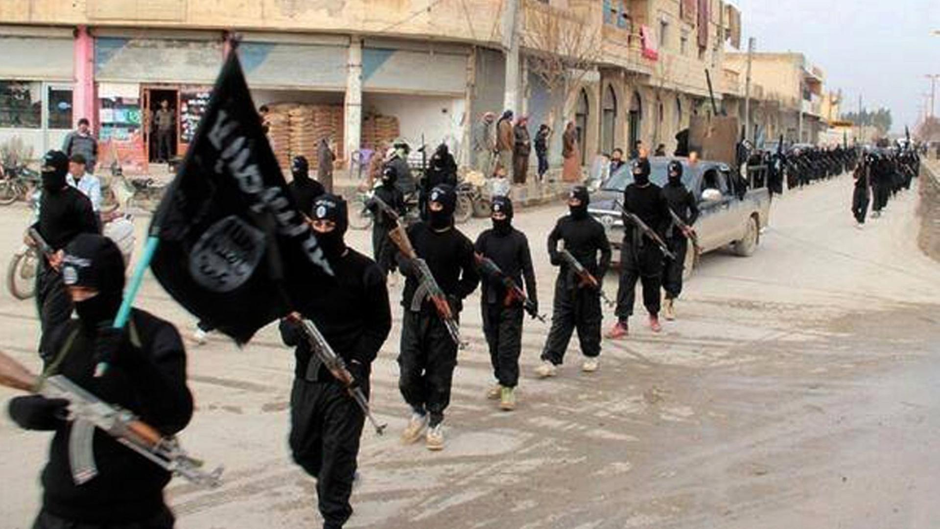 Ein Propagandabild der Terrormiliz IS: Mitglieder in schwarzen Overalls und mit Gewehren in der Hand marschieren durch Raqqa in Syrien. Der erste Mann trägt die schwarz IS-Fahne. Ein Propagandabild der Terrormiliz IS: Mitglieder in schwarzen Overalls und mit Gewehren in der Hand marschieren durch Raqqa in Syrien. Der erste Mann trägt die schwarz IS-Fahne.