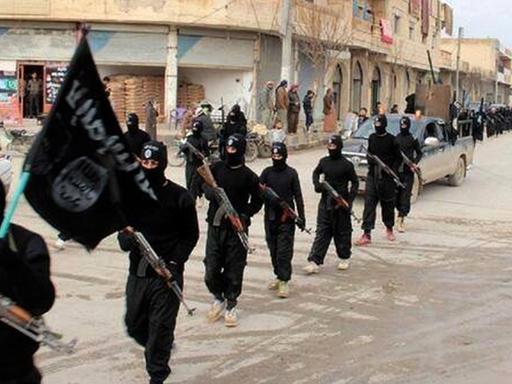 Ein Propagandabild der Terrormiliz IS: Mitglieder in schwarzen Overalls und mit Gewehren in der Hand marschieren durch Raqqa in Syrien. Der erste Mann trägt die schwarz IS-Fahne.