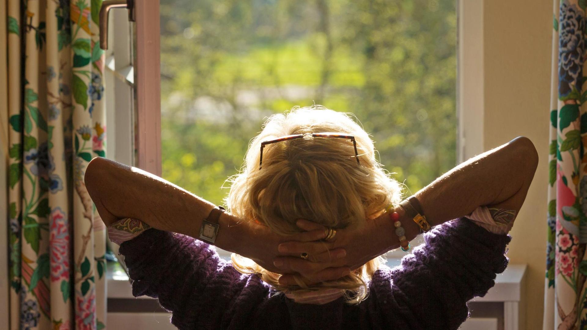 Ein Frau sitzt, mit im Nacken verschränkten Armen, vor einem offenen Fenster. Ein Frau sitzt, mit im Nacken verschränkten Armen, vor einem offenen Fenster.