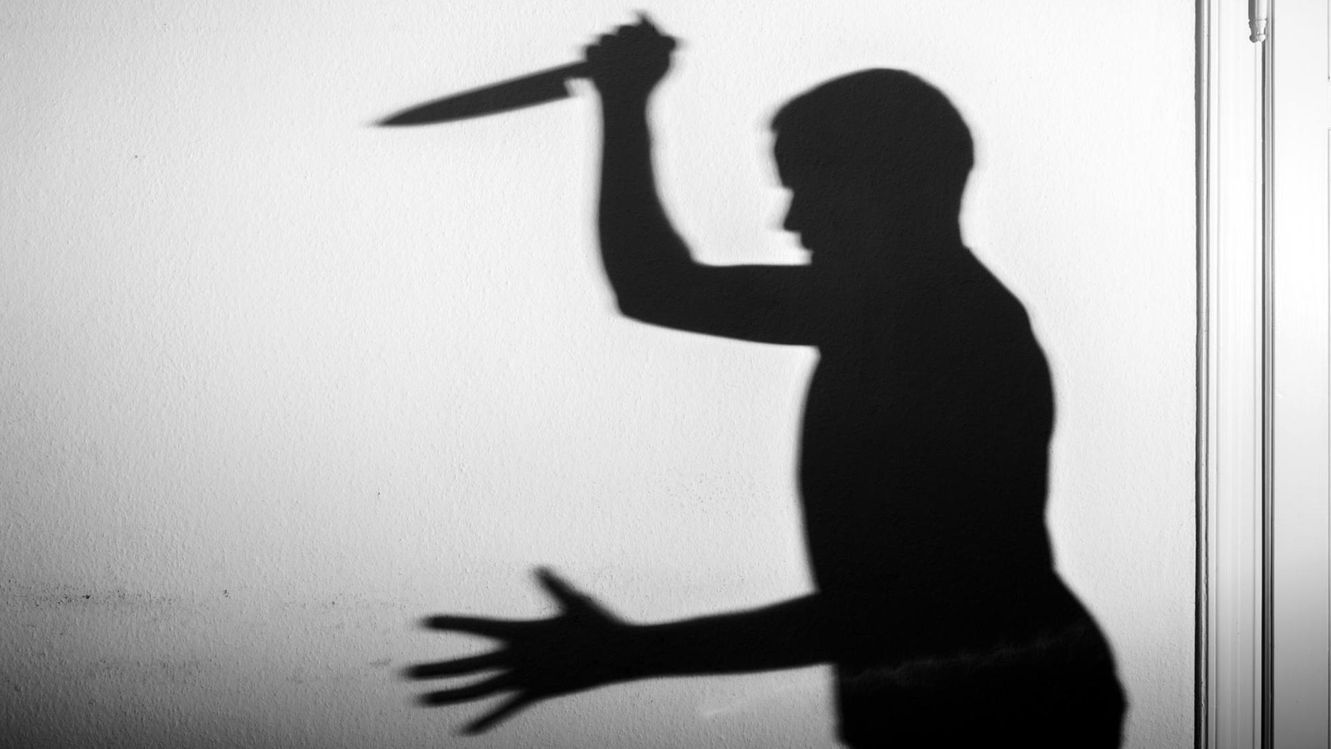 Der Schatten eines Mannes mit einem großen Küchenmesser in der Hand wird an eine weiße Wand geworfen.