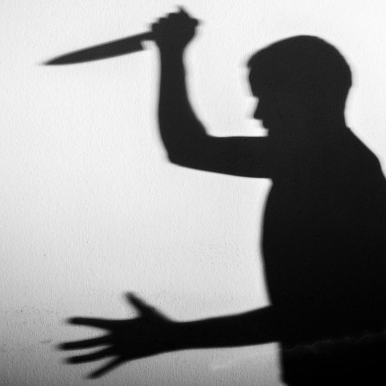 Der Schatten eines Mannes mit einem großen Küchenmesser in der Hand wird an eine weiße Wand geworfen.