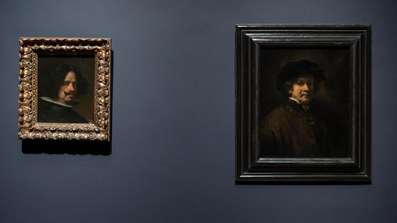 Blick in die Austellung. Zwei Selbstportäts von Rembrandt und Velázquez hängen nebeneinander an einer dunkelblauen Wand. Blick in die Austellung. Zwei Selbstportäts von Rembrandt und Velázquez hängen nebeneinander an einer dunkelblauen Wand.