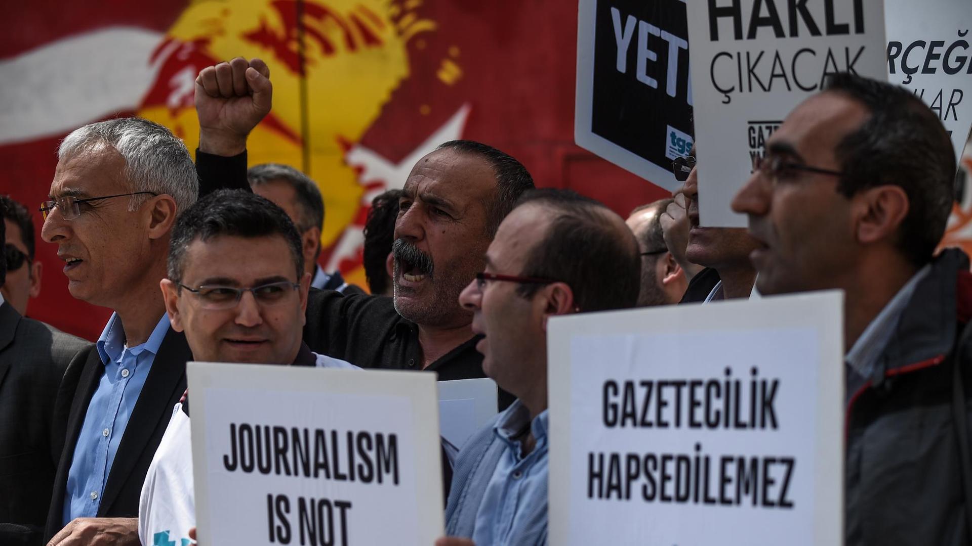 Mehrere Männer stehen in einer Menge, einige halten Plakate mit der Aufschrift "Journalismus ist kein Verbrechen".