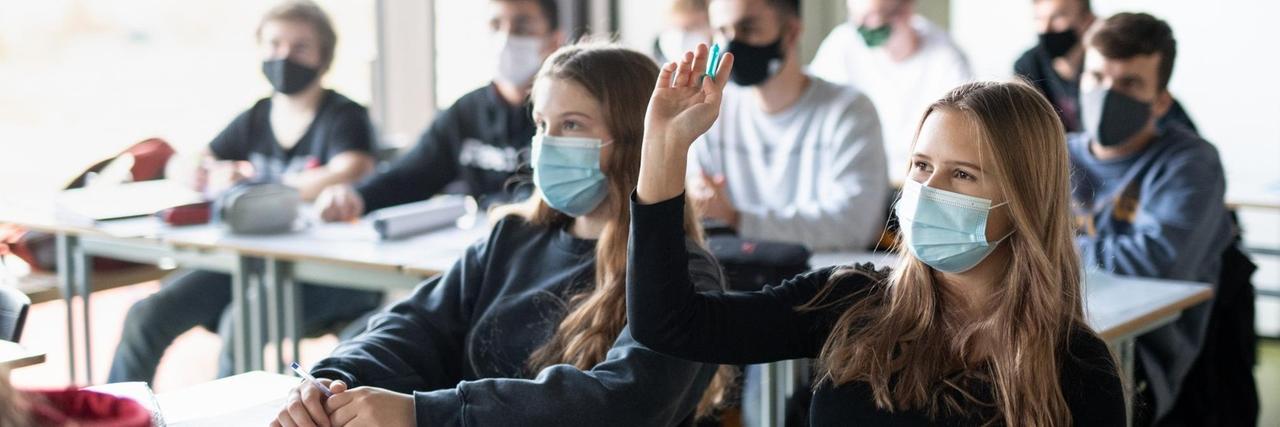 Schülerinnen und Schüler nehmen mit Mund- und Nasenschutz am Unterricht teil, eine Schülerin meldet sich. Schülerinnen und Schüler nehmen mit Mund- und Nasenschutz am Unterricht teil, eine Schülerin meldet sich.