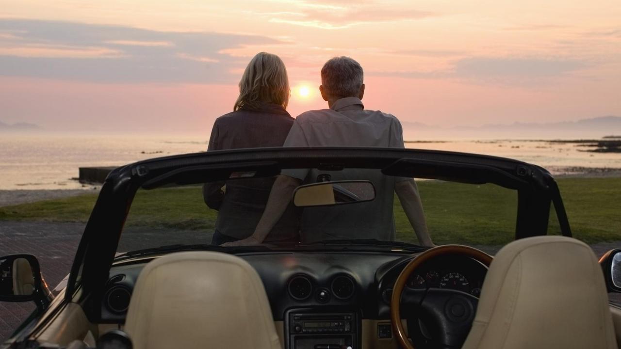 Ein Paar lehnt sich an ein Cabrio und bewundert den Sonnenuntergang am Meer.