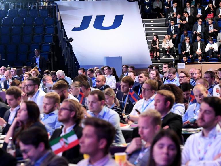 Blick von vorne auf die Delegierten in der Halle; im Hintergrund ein weißes "JU"-Banner.