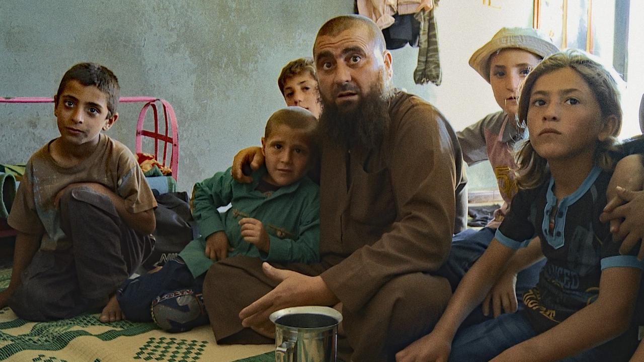 Ein Mann mit Bart und kahl geschorenem Kopf sitzt von Kindern umringt auf einer Decke in einem kargen Haus. Alle schauen aufmerksam in Richtung des Betrachters. Ein Mann mit Bart und kahl geschorenem Kopf sitzt von Kindern umringt auf einer Decke in einem kargen Haus. Alle schauen aufmerksam in Richtung des Betrachters.