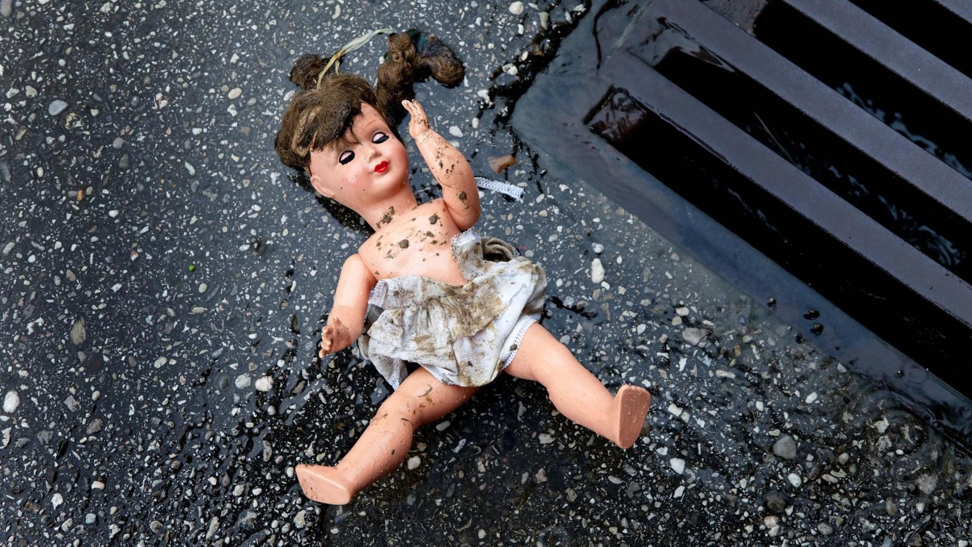 Zu sehen ist eine Puppe mit schmutzigem Kleid, die auf dem Boden neben einem Gulli liegt. Zu sehen ist eine Puppe mit schmutzigem Kleid, die auf dem Boden neben einem Gulli liegt.