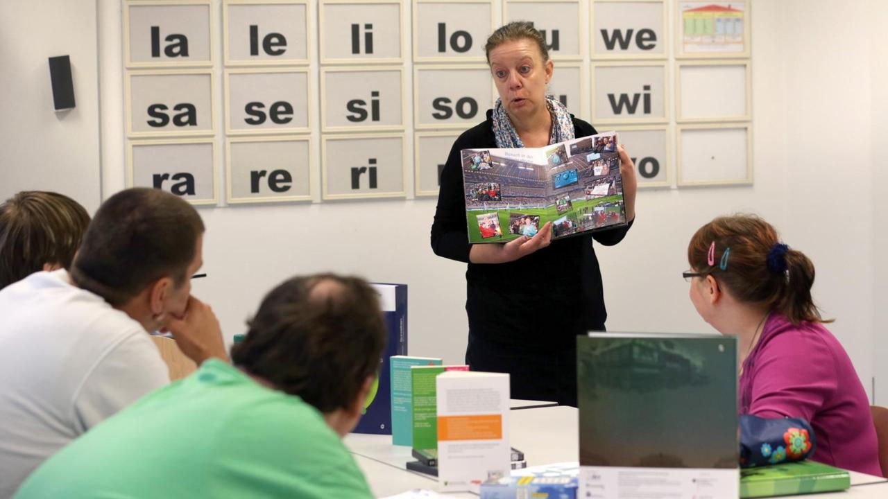 Erwachsene sitzen in einem Klassenzimmer. Eine Lehrerin zeigt ein Buch. An der Tafel sind Buchstaben zu lesen.