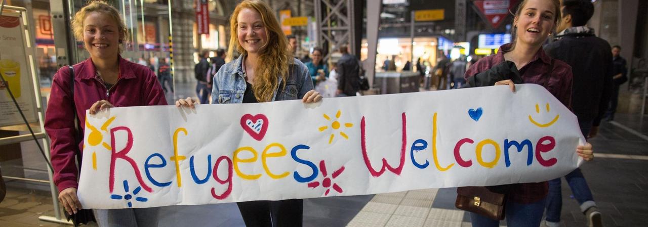 Drei junge Frauen stehen mit einem Begrüßungsplakat mit der Aufschrift "Refugees Welcome" für Flüchtlinge an den Gleisen.
