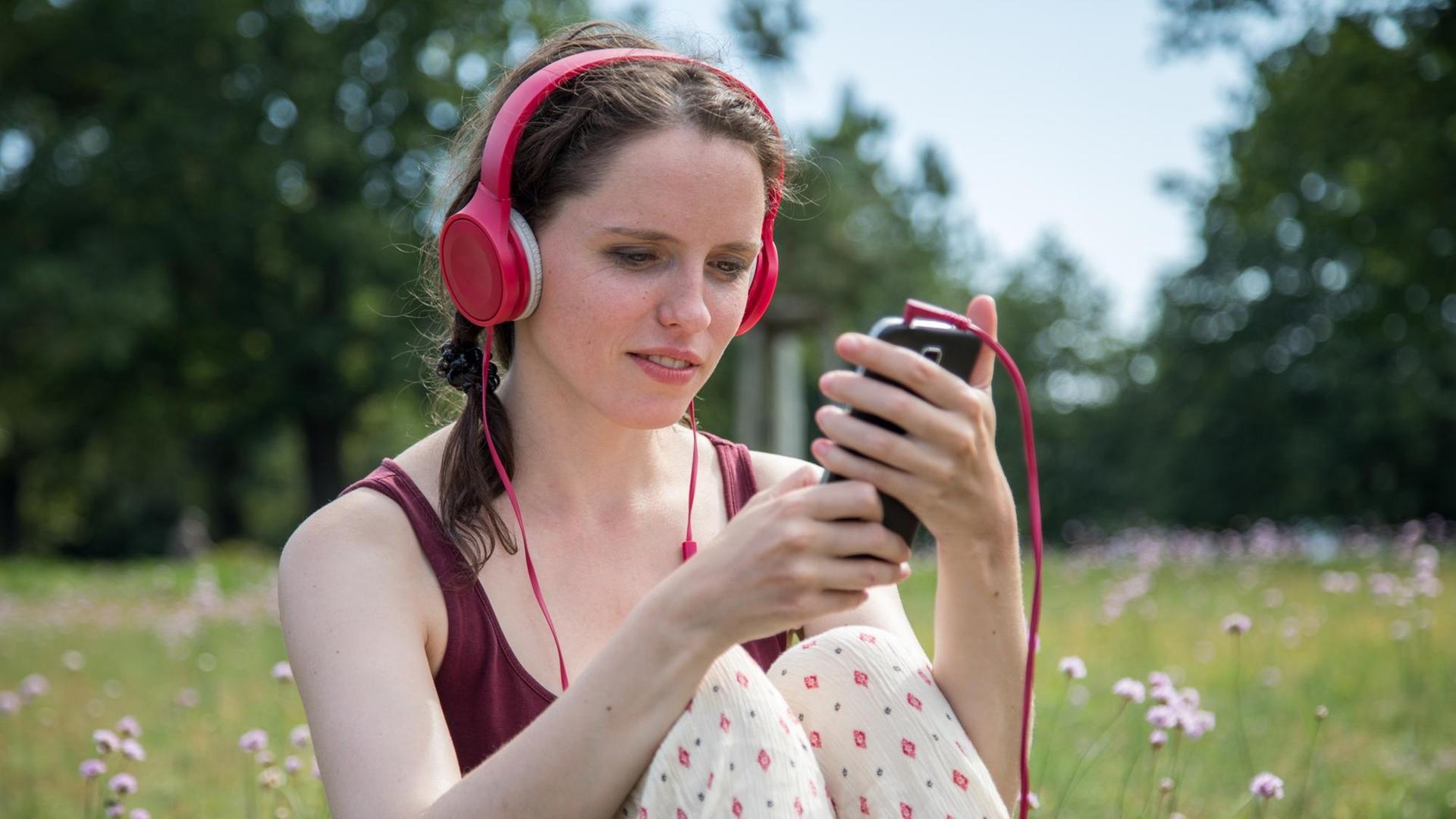 Eine Frau sitzt an einem Badesee in Oranienburg (Brandenburg) auf einer Wiese und hört Musik mit ihrem Handy. Eine Frau sitzt an einem Badesee in Oranienburg (Brandenburg) auf einer Wiese und hört Musik mit ihrem Handy.