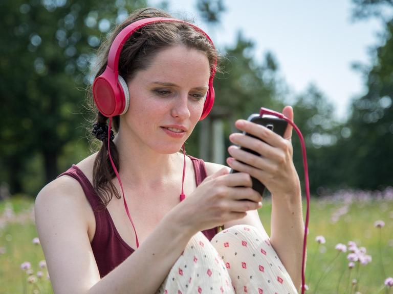 Eine Frau sitzt an einem Badesee in Oranienburg (Brandenburg) auf einer Wiese und hört Musik mit ihrem Handy.