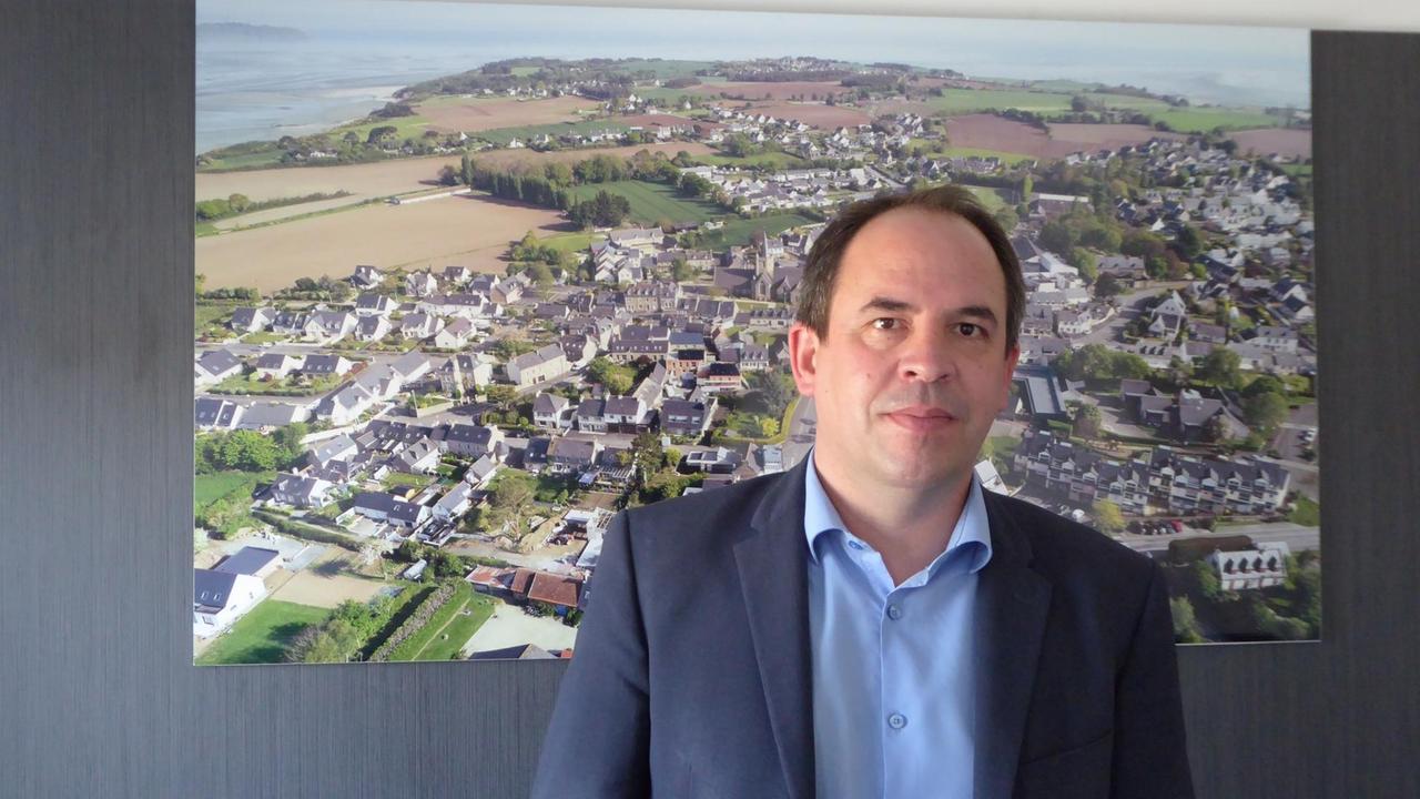 Ein Mann mit Halbglatze, Jacket und hellblauem Hemd steht vor einem großen Wandfoto, auf dem eine Küstenlandschaft abgebildet ist. Ein Mann mit Halbglatze, Jacket und hellblauem Hemd steht vor einem großen Wandfoto, auf dem eine Küstenlandschaft abgebildet ist.