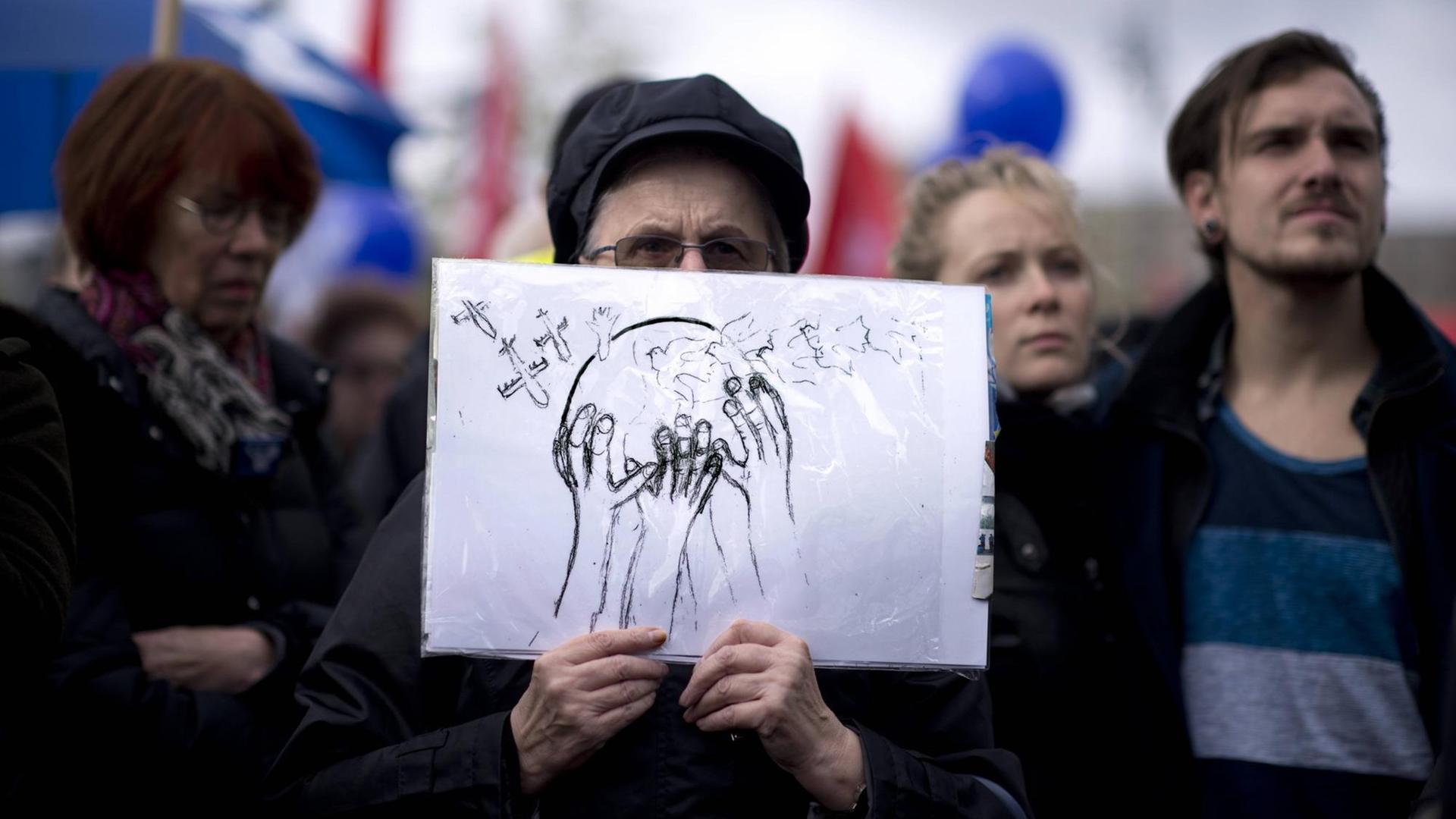 Ein Demonstrant auf einer Friedensdemo hält ein Schild vor sich. Auf dem Schild ist eine Weltkugel abgebildet die von mehreren Händen festgehalten wird. Auf die Kugeln fliegen von der einen Seite Friedenstauben und von der anderen Kampfflugzeuge zu. 