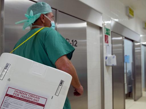Ein Mann in grüner OP-Kleidung trägt einen Styropor-Behälter für den Transport von Spenderorganen an einem Operationssaal vorbei.