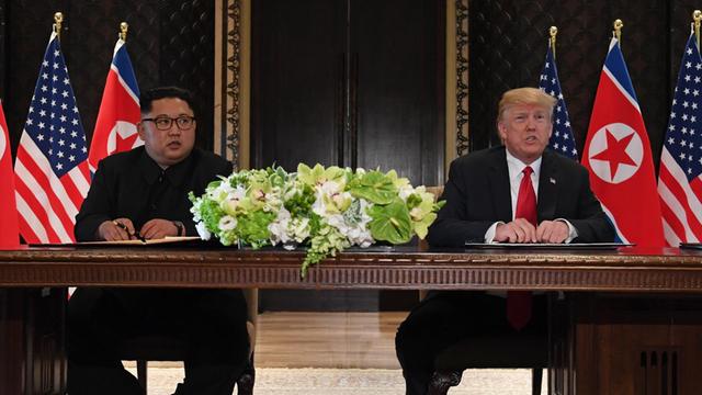 Kim (l.) und Trump sitzen nebeneinander an einem Tisch, vor sich jeweils eine Dokumentenmappe. Im Hintergrund die Nationalflaggen.