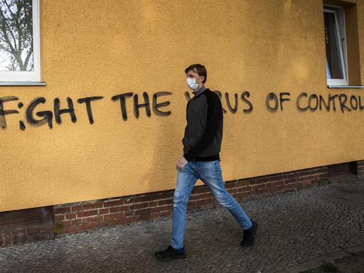 Ein Mann geht in Berlin an einer dunkelgelben Hauswand entlang, auf der in schwarzen Großbuchstaben gesprüht ist: 'Fight the virus of control' (zu Deutsch: Kampf dem Virus der Kontrolle)