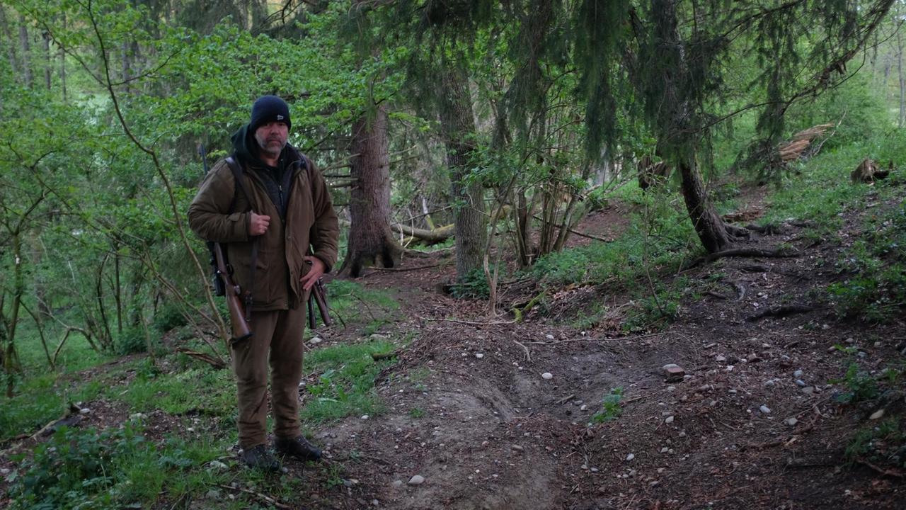 Ein Mann in Jagdkleidung mit dem Gewehr über der Schulter geht durch einen Wald.