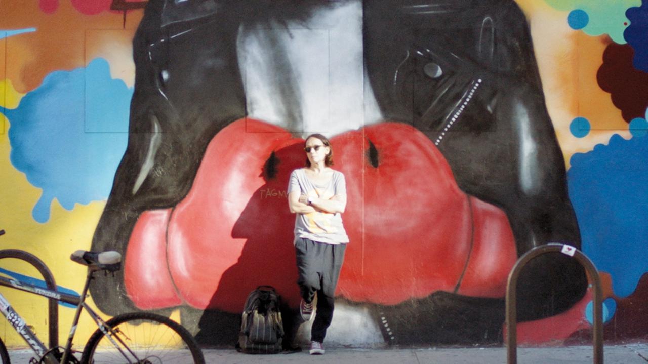 Marion Brasch lehnt an einer Wand mit einem Boxhandschuh-Graffiti in einer Szene aus dem Film "Familie Brasch"