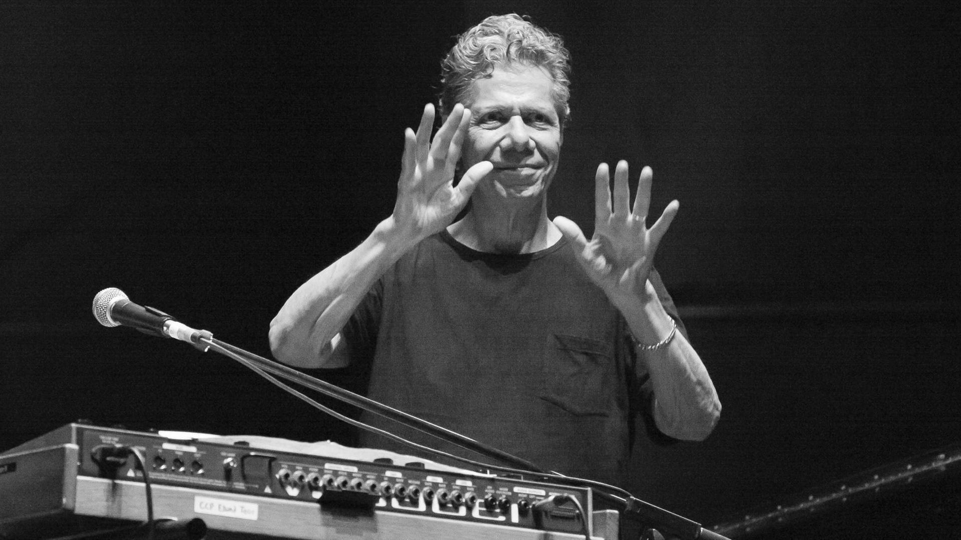 Die Schwarzweißfotografie zeigt einen Mann im T-Shirt mit grauem, weilligen Haar hinter einem E-Piano. Er hält beide Hände in die Höhe, die Handflächen zeigen wie zum Segen nach vorn. Die Schwarzweißfotografie zeigt einen Mann im T-Shirt mit grauem, weilligen Haar hinter einem E-Piano. Er hält beide Hände in die Höhe, die Handflächen zeigen wie zum Segen nach vorn.