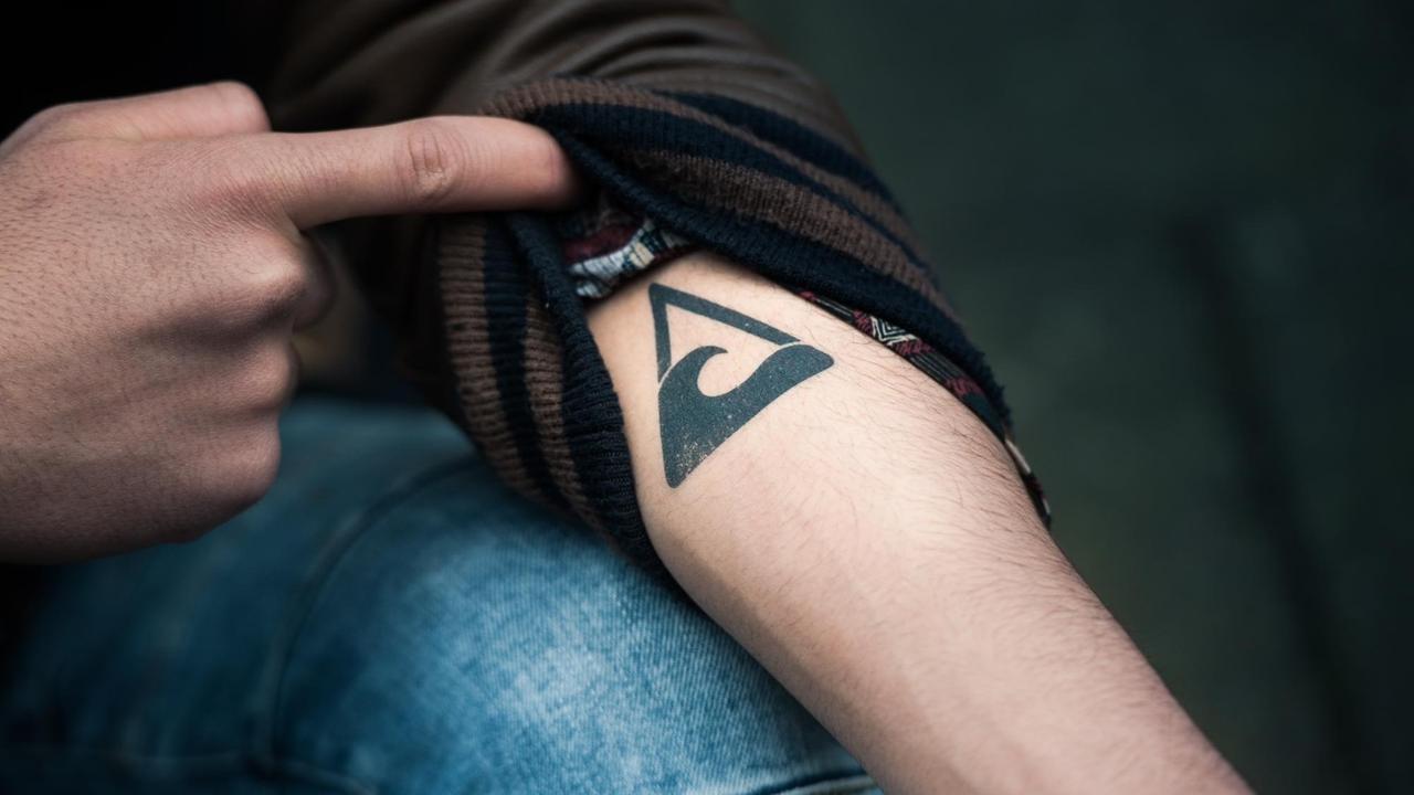 Ein Unterarm, auf dem ein Wellensymbol in einem Dreieck tätowiert ist. Ein Unterarm, auf dem ein Wellensymbol in einem Dreieck tätowiert ist.
