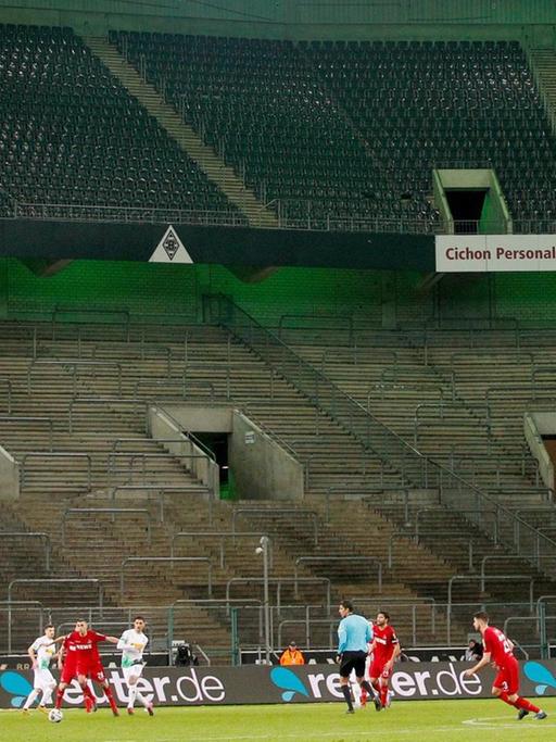 Borussia Mönchengladbach und der 1. FC Köln spielen im Borussia-Park vor leeren Zuschauerrängen. Im Vordergrund spielen sie Mannschaften gegeneinander. Im Hintergrund sind sie leeren Ränge zu sehen.