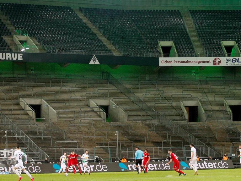 Borussia Mönchengladbach und der 1. FC Köln spielen im Borussia-Park vor leeren Zuschauerrängen. Im Vordergrund spielen sie Mannschaften gegeneinander. Im Hintergrund sind sie leeren Ränge zu sehen.