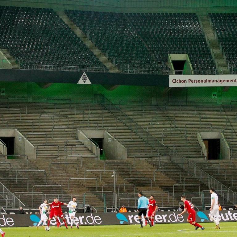 Borussia Mönchengladbach und der 1. FC Köln spielen im Borussia-Park vor leeren Zuschauerrängen. Im Vordergrund spielen sie Mannschaften gegeneinander. Im Hintergrund sind sie leeren Ränge zu sehen.