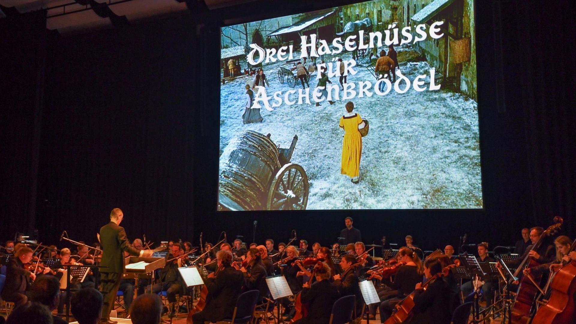 Auf eine Leinwand wird der Film "Drei Haselnüsse für Aschenbrödel" projiziert, davor sitzt ein Orchester und spielt die Filmmusik live. Auf eine Leinwand wird der Film "Drei Haselnüsse für Aschenbrödel" projiziert, davor sitzt ein Orchester und spielt die Filmmusik live.