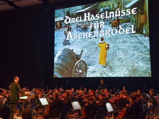 Auf eine Leinwand wird der Film "Drei Haselnüsse für Aschenbrödel" projiziert, davor sitzt ein Orchester und spielt die Filmmusik live.