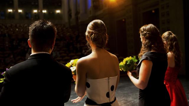 Junge Sänger und Sängerinnen stehen auf der Bühne mit Blumensträußen, die der Kamera den Rücken zuwenden, weil sie sich beim Publikum für den Beifall bedenken.
