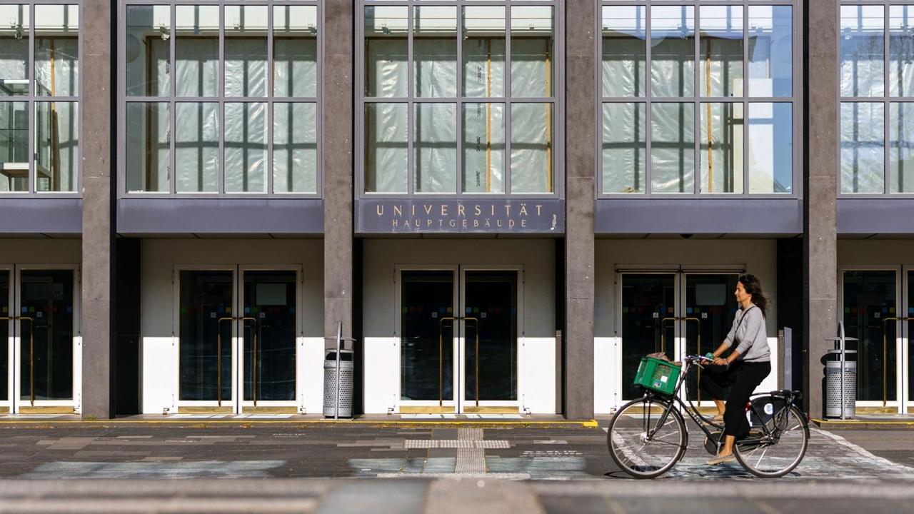 Der fast menschenleere Eingang der Albertus Magnus Universität zu Köln, eine Fahrradfahrerin fährt vorbei.
