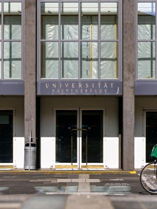 Der fast menschenleere Eingang der Albertus Magnus Universität zu Köln, eine Fahrradfahrerin fährt vorbei.