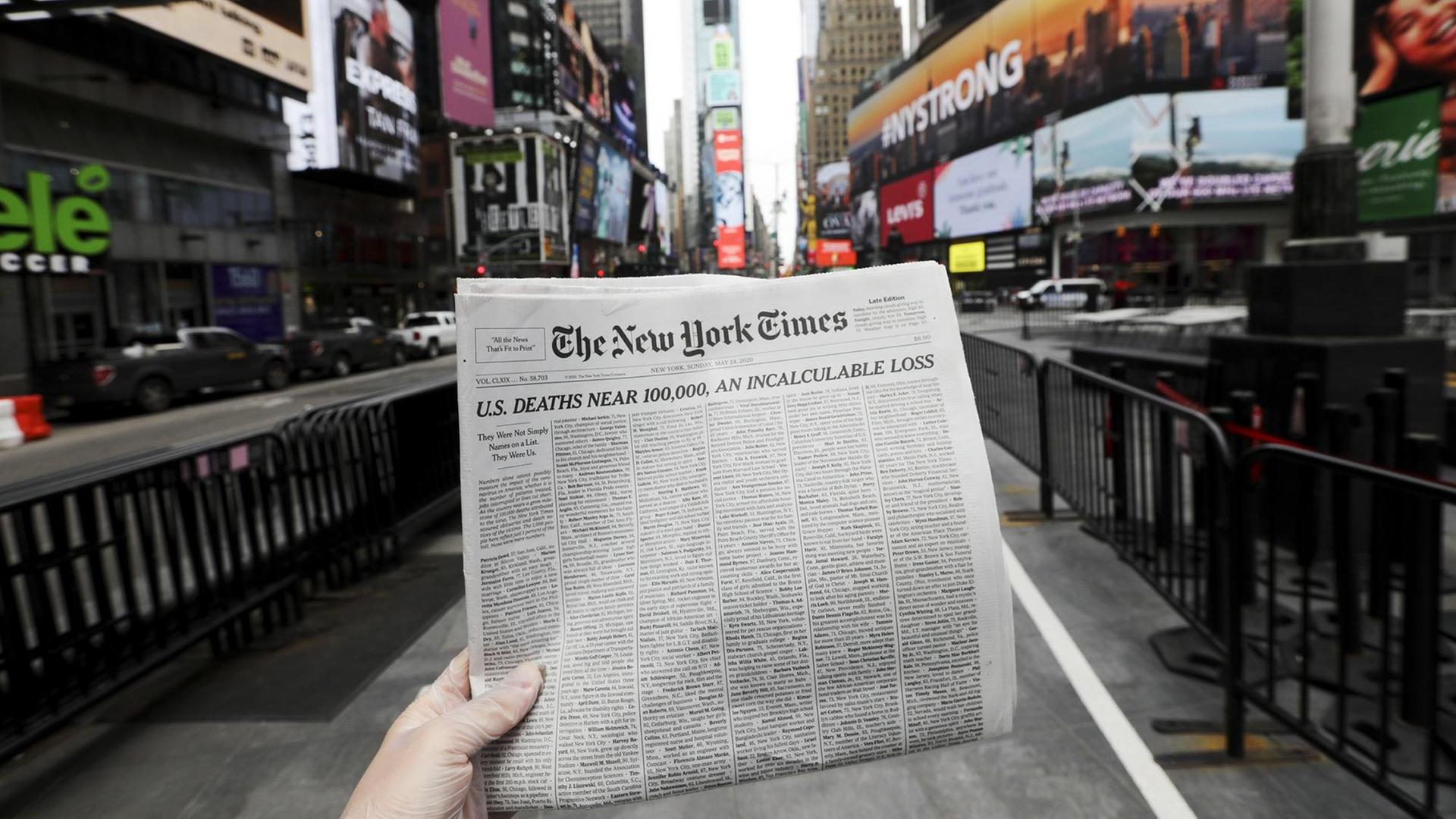 Eine Händ hält die Titelseite der New York Times, vor die Kulisse der Time Square in New York.