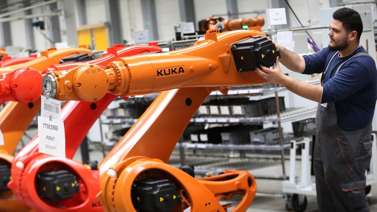 Ein Arbeiter hantiert in einer Halle an einem orangefarbenen Kuka-Roboter, daneben stehen weitere Roboter der gleichen Bauart. Ein Arbeiter hantiert in einer Halle an einem orangefarbenen Kuka-Roboter, daneben stehen weitere Roboter der gleichen Bauart.