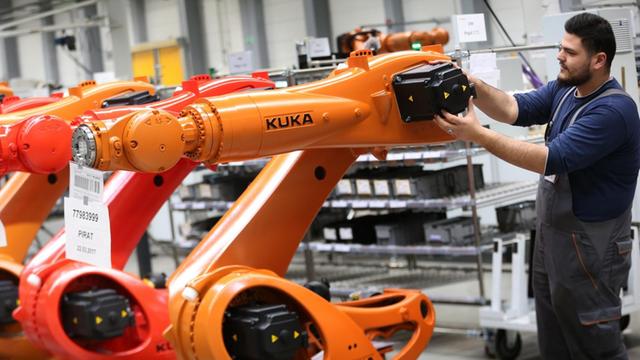 Ein Arbeiter hantiert in einer Halle an einem orangefarbenen Kuka-Roboter, daneben stehen weitere Roboter der gleichen Bauart.