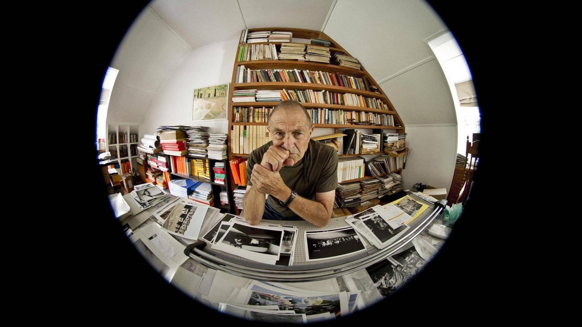 Der südafrikanische Fotograf David Goldblatt sitzt in seinem Büro in Johannesburg am Schreibtisch, auf dem Schwarz-Weiß-Abzüge von Fotos liegen. Hinter ihm steht eine Bücherwand. Goldblatt hat beide Ellbogen aufgestützt und blickt nachdenklich in die Kamera. Das Foto ist rund und wurde in Fischaugen-Perspektive aufgenommen. Der südafrikanische Fotograf David Goldblatt sitzt in seinem Büro in Johannesburg am Schreibtisch, auf dem Schwarz-Weiß-Abzüge von Fotos liegen. Hinter ihm steht eine Bücherwand. Goldblatt hat beide Ellbogen aufgestützt und blickt nachdenklich in die Kamera. Das Foto ist rund und wurde in Fischaugen-Perspektive aufgenommen.
