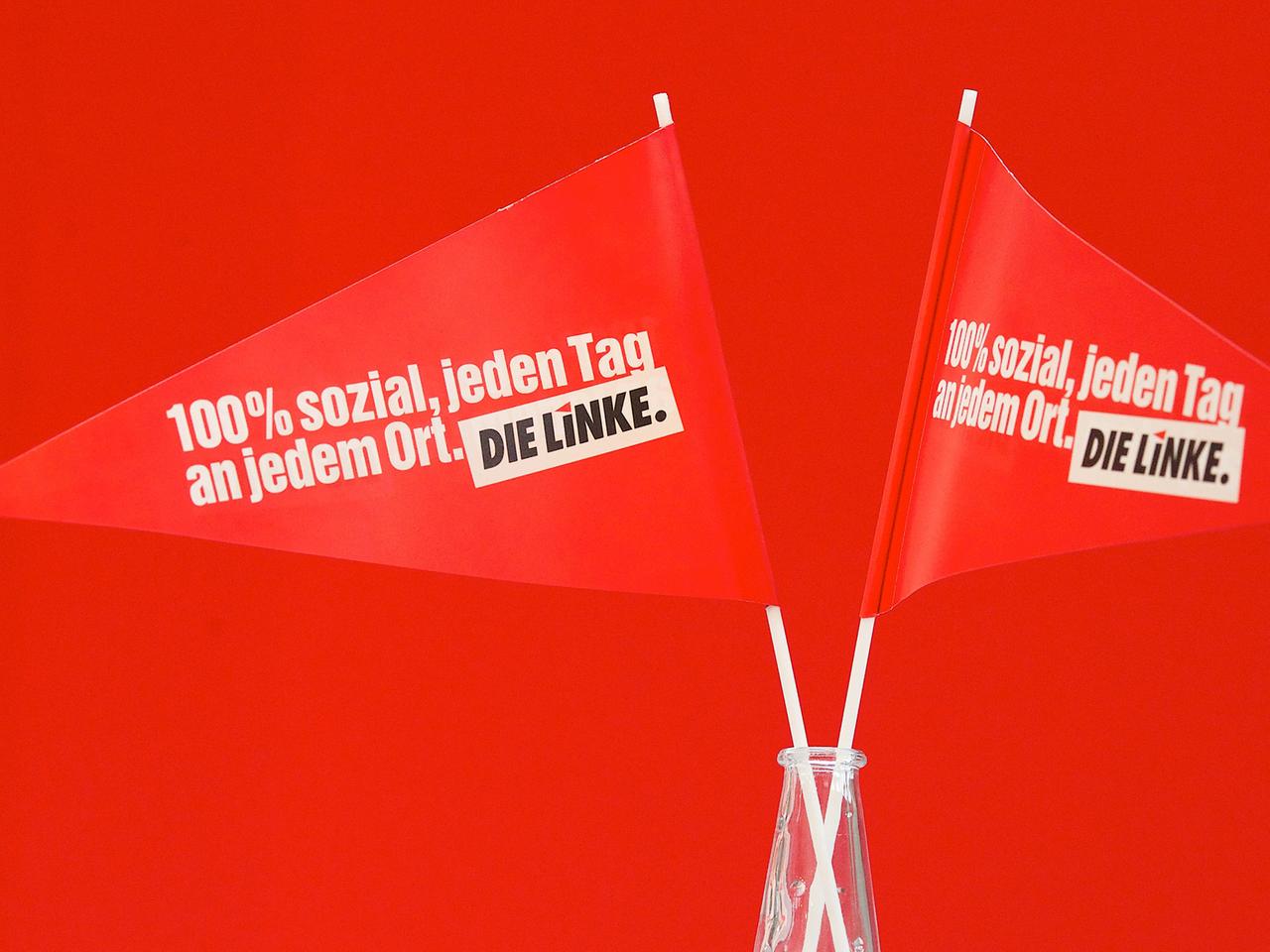 Zwei rote Fähnchen stehen vor rotem Hintergrund in einem Glas. Aufschrift: 100 Prozent sozial, jeden Tag, an jedem Ort. Die Linke.