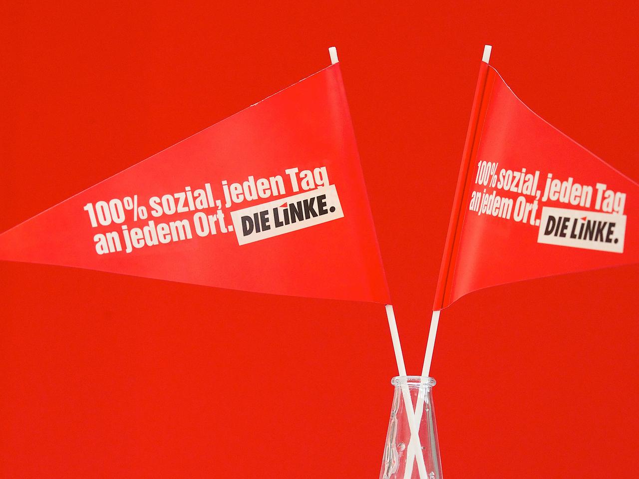 Zwei rote Fähnchen stehen vor rotem Hintergrund in einem Glas. Aufschrift: 100 Prozent sozial, jeden Tag, an jedem Ort. Die Linke. Zwei rote Fähnchen stehen vor rotem Hintergrund in einem Glas. Aufschrift: 100 Prozent sozial, jeden Tag, an jedem Ort. Die Linke.